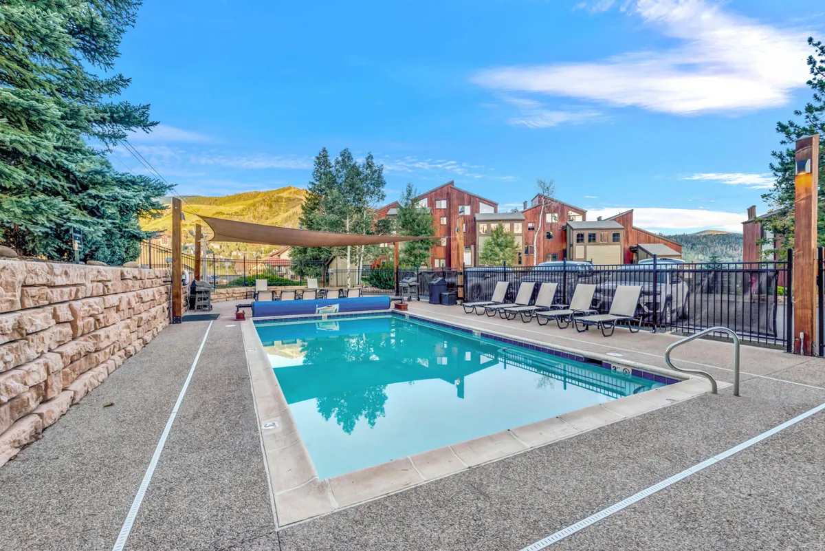 A resort-style swimming pool surrounded by lounge chairs, lush greenery, and a mountain backdrop. The area invites guests to unwind, socialize, or soak up the sun, reflecting the amenities available for long-term stays.