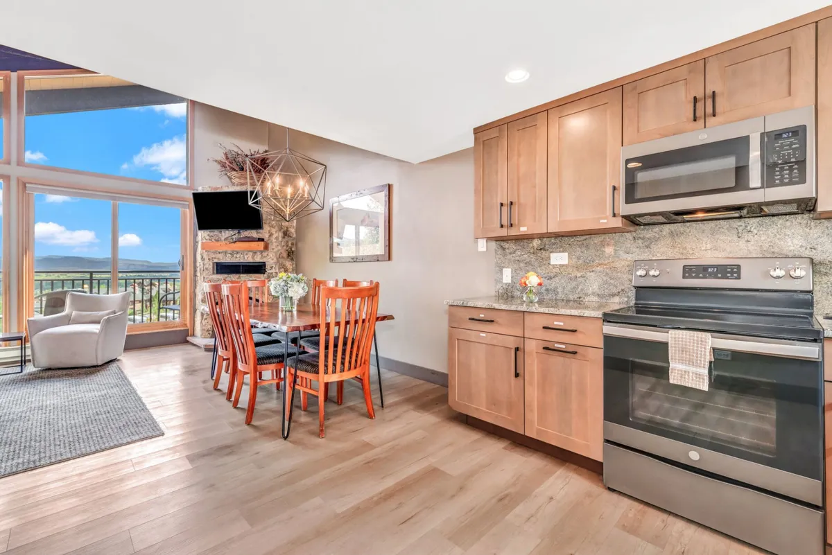 A spacious, welcoming area is depicted with a large dining table surrounded by chairs, seamlessly connected to the kitchen. Large windows flood the space with light and offer scenic views, while the living area nearby promises comfort and further opportunities for socializing.