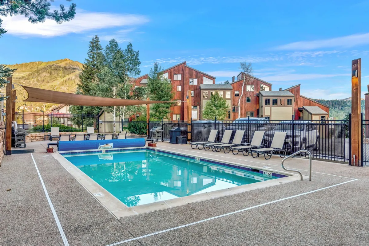 A vibrant exterior scene featuring a sparkling swimming pool surrounded by lounge chairs and a shade sail, with a dramatic mountain backdrop. The landscaping adds to the inviting, resort-like feel, suggesting a perfect spot for family pool days or gatherings.