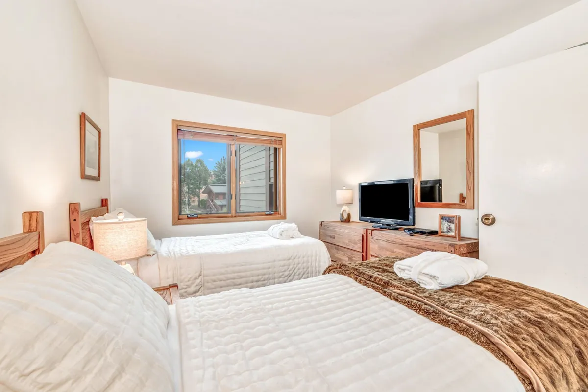 A welcoming second bedroom with two twin beds, a wooden dresser, window with a view, television, and a wall mirror. The layout is ideal for kids or friends, offering both entertainment and relaxation.