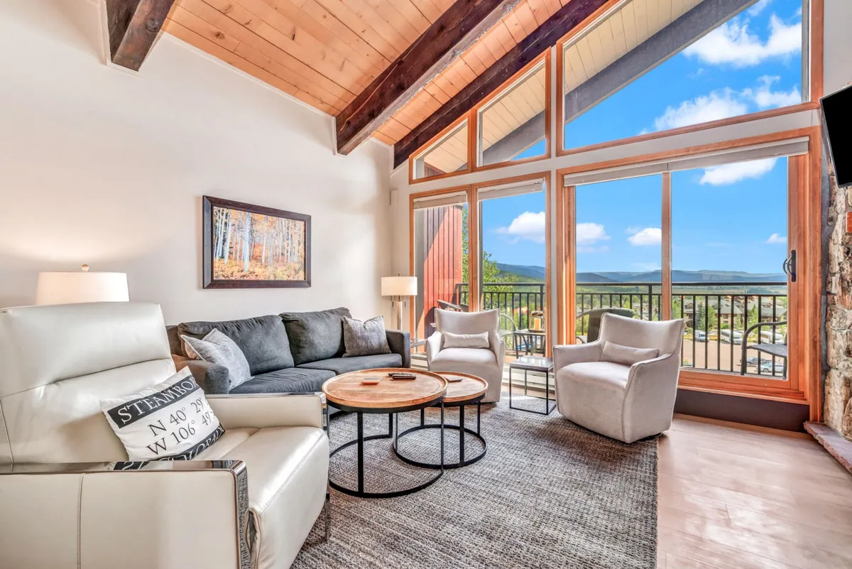The image shows a stylishly updated condo interior featuring inviting, plush seating arranged around a wooden coffee table. Large windows flood the space with natural light and frame picturesque mountain views. Exposed wooden beams add warmth and character, and tasteful artwork adorns the walls, enhancing the welcoming ambiance.
