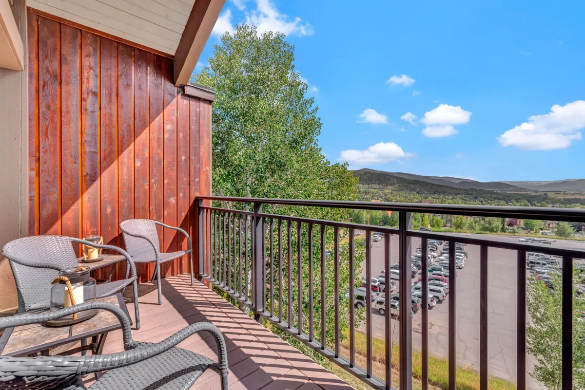 This image features a balcony with a railing, two chairs, a table with drinks, green trees, and a stunning mountain view. It captures the essence of relaxing outdoors while enjoying beautiful scenery.