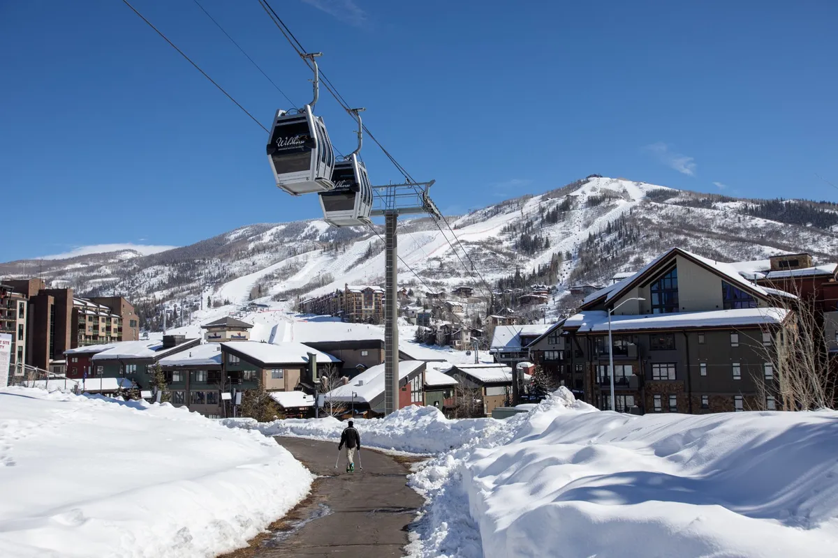 A vibrant winter scene featuring a cable car, snow-covered buildings, and a skier walking through the heart of the Steamboat resort village. The landscape is blanketed in snow, with mountains rising in the background and the lively atmosphere of the base area visible.