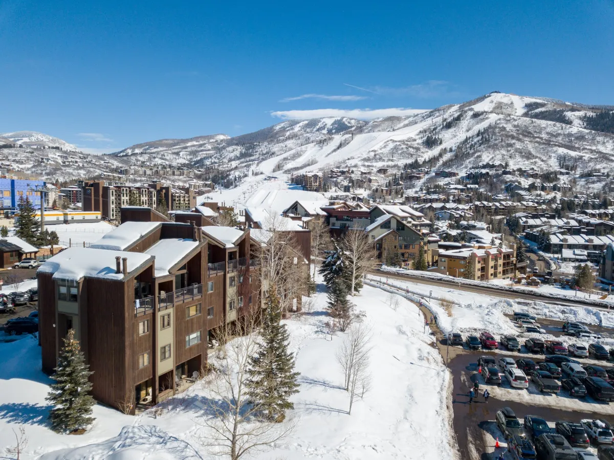 A panoramic view showcasing snow-covered mountains, active ski slopes, and the charming wooden architecture of nearby condos. The scene also includes a parking area and trees, reinforcing the mountain resort ambiance.