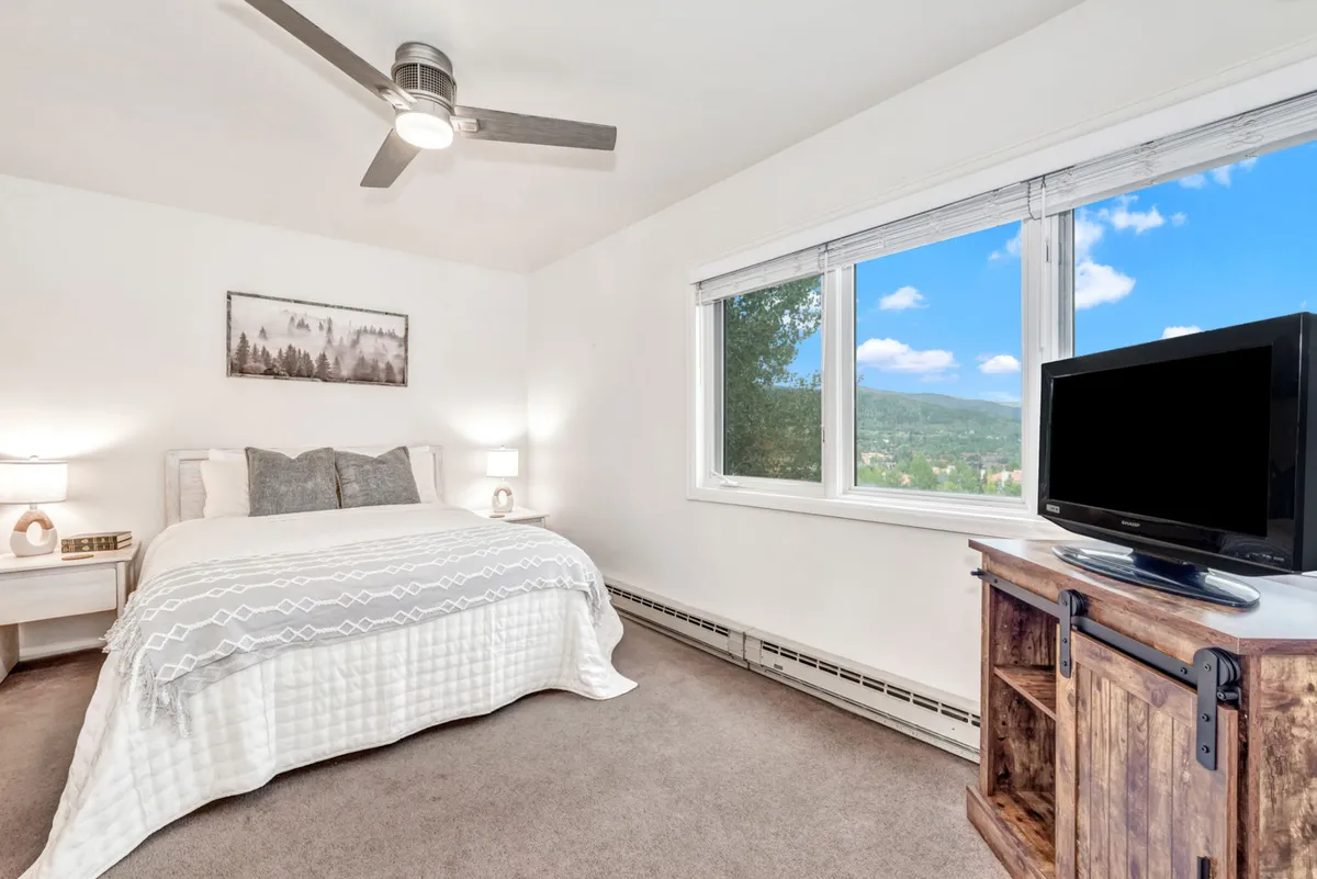 A serene primary bedroom with a decorative bedspread, a large window offering a scenic view, and a wooden TV stand at the foot of the bed. The space exudes comfort and has a peaceful atmosphere—ideal for unwinding.