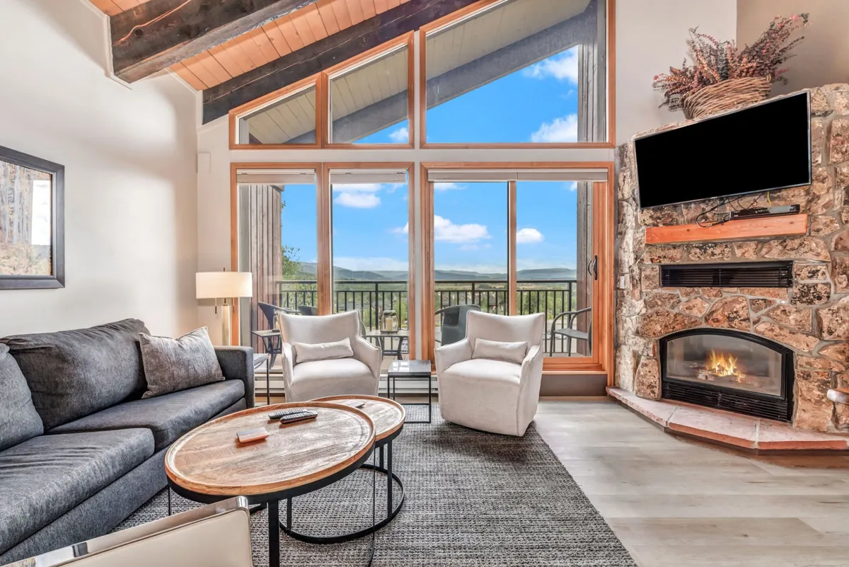 This image showcases a cozy, open living space with a large stone fireplace, comfortable seating, a round coffee table, and expansive windows revealing a dramatic mountain view. The seating arrangement is oriented to encourage group gatherings and conversation, while the windows flood the room with natural light and connect the interior to the surrounding alpine scenery.