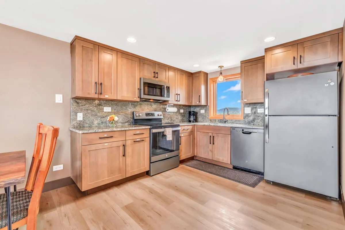 This image presents a bright and inviting kitchen with modern stainless steel appliances, warm wooden cabinetry, and abundant natural light streaming in from a window. The focal point is the functional workspace highlighted by granite countertops, with enough space for group meal prep or casual dining.